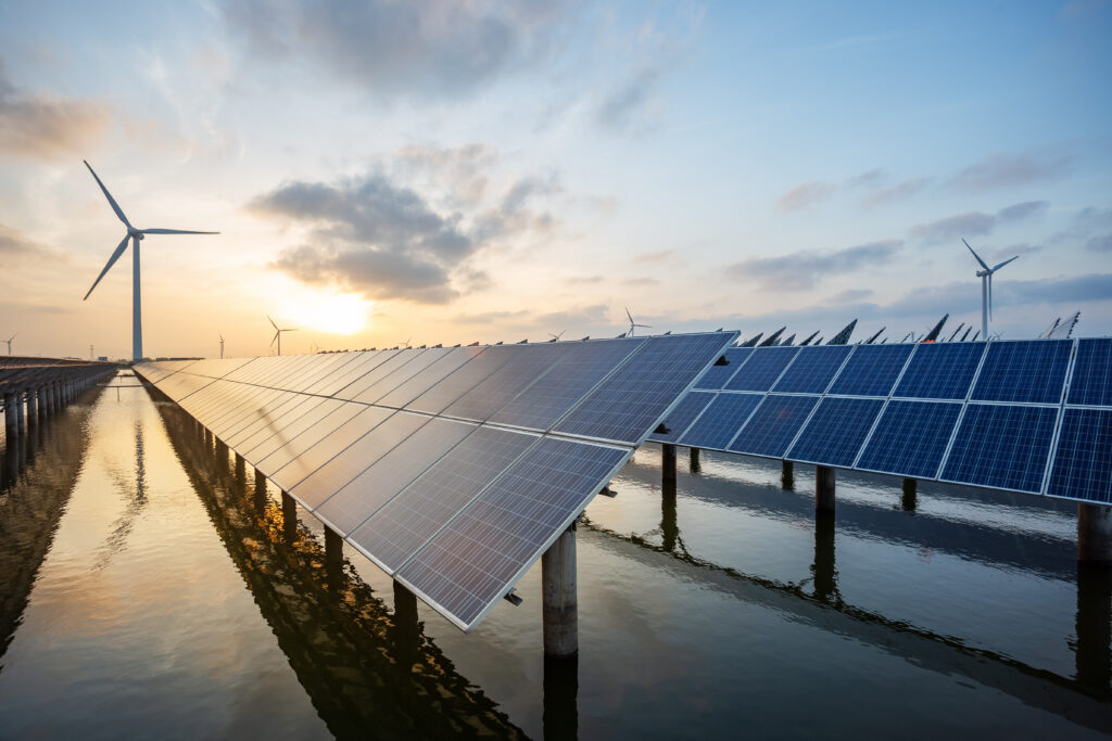 A picturesque sunset or sunrise backdrop with stunning hues of orange, pink, and purple. In the foreground, rows of solar panels stand tall, gleaming under the soft sunlight. In the distance, majestic wind turbines generate clean energy against the vibrant sky, symbolizing the harmonious coexistence of renewable energy sources and nature's beauty.